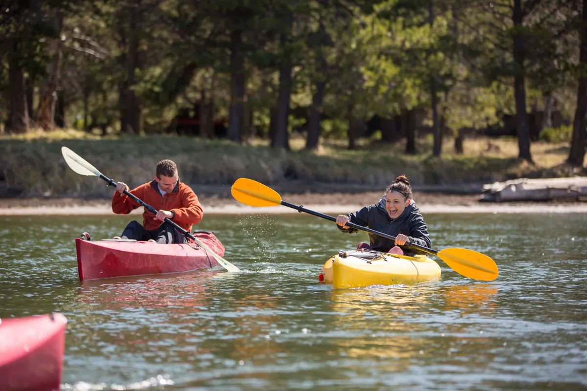 Two kayakers in vibrant kayaks paddle across calm water, their reflections mirrored on the surface. The scene radiates energy and togetherness, capturing the essence of shared outdoor experiences on the river.