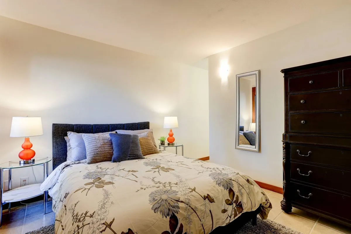 A cozy and tranquil bedroom with a comfortable bed dressed in floral bedding, flanked by nightstands with lamps. A tall dresser and a wall mirror add to the room’s functionality and charm, with gentle daylight enhancing the peaceful mood.