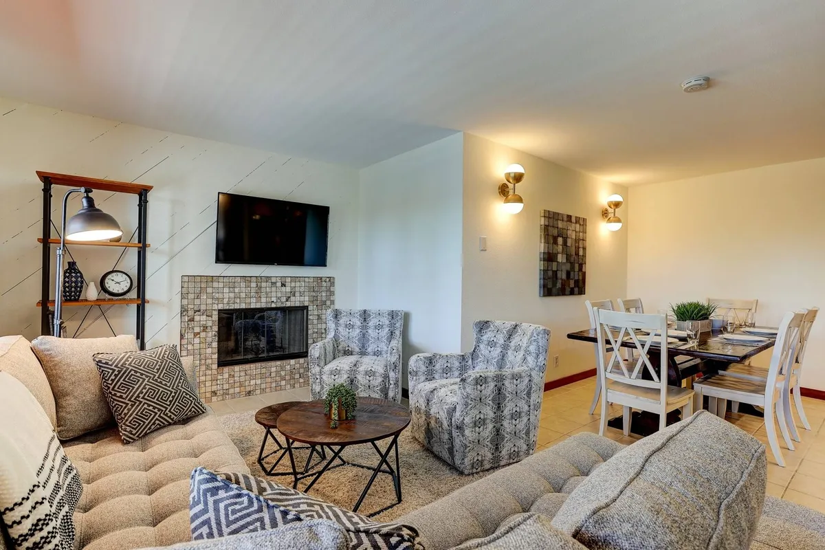A cozy, inviting interior with a plush sofa and fireplace, a nearby dining table, decorative shelving, and wall art. The space is warm and welcoming, suited for family gatherings, relaxing evenings, and shared meals after a day of activity.
