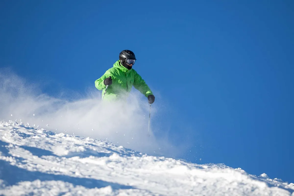 A skier in a bright green jacket makes their way down a sunlit, snowy mountain slope under a clear blue sky, with expansive views of the landscape. The energy and excitement of the moment are palpable, suggesting both adventure and natural beauty.