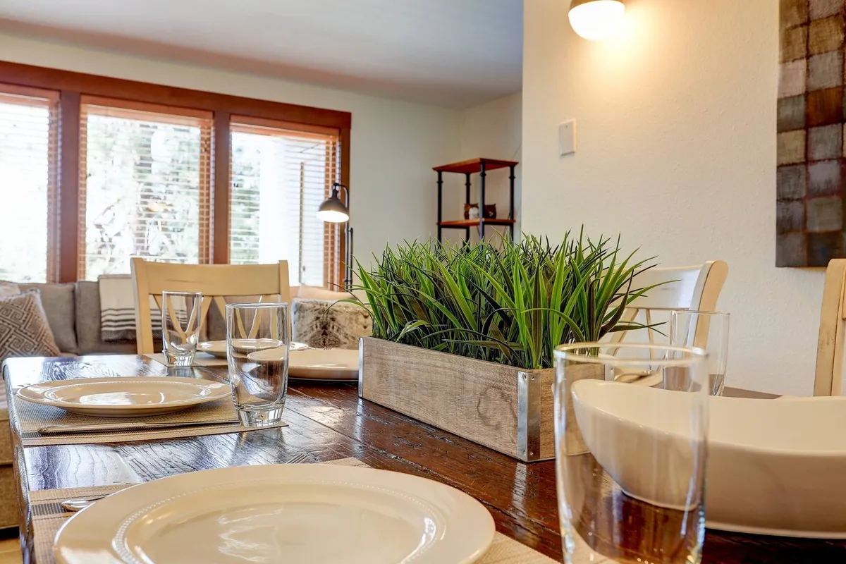 The image shows a welcoming dining area with a table set for meals, a lush greenery centerpiece, and a large window that provides glimpses of the outdoors. The space is filled with natural light, creating a connection between the indoor setting and the surrounding environment.