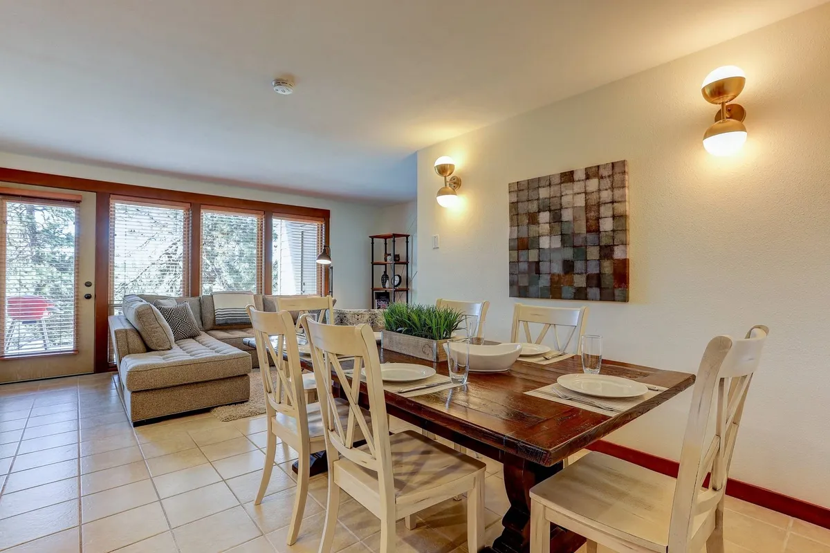A bright, welcoming dining area with a large table, comfortable sofa, and oversized windows letting in natural light. It’s set up for family dining, games, and socializing, with tasteful artwork on the walls.