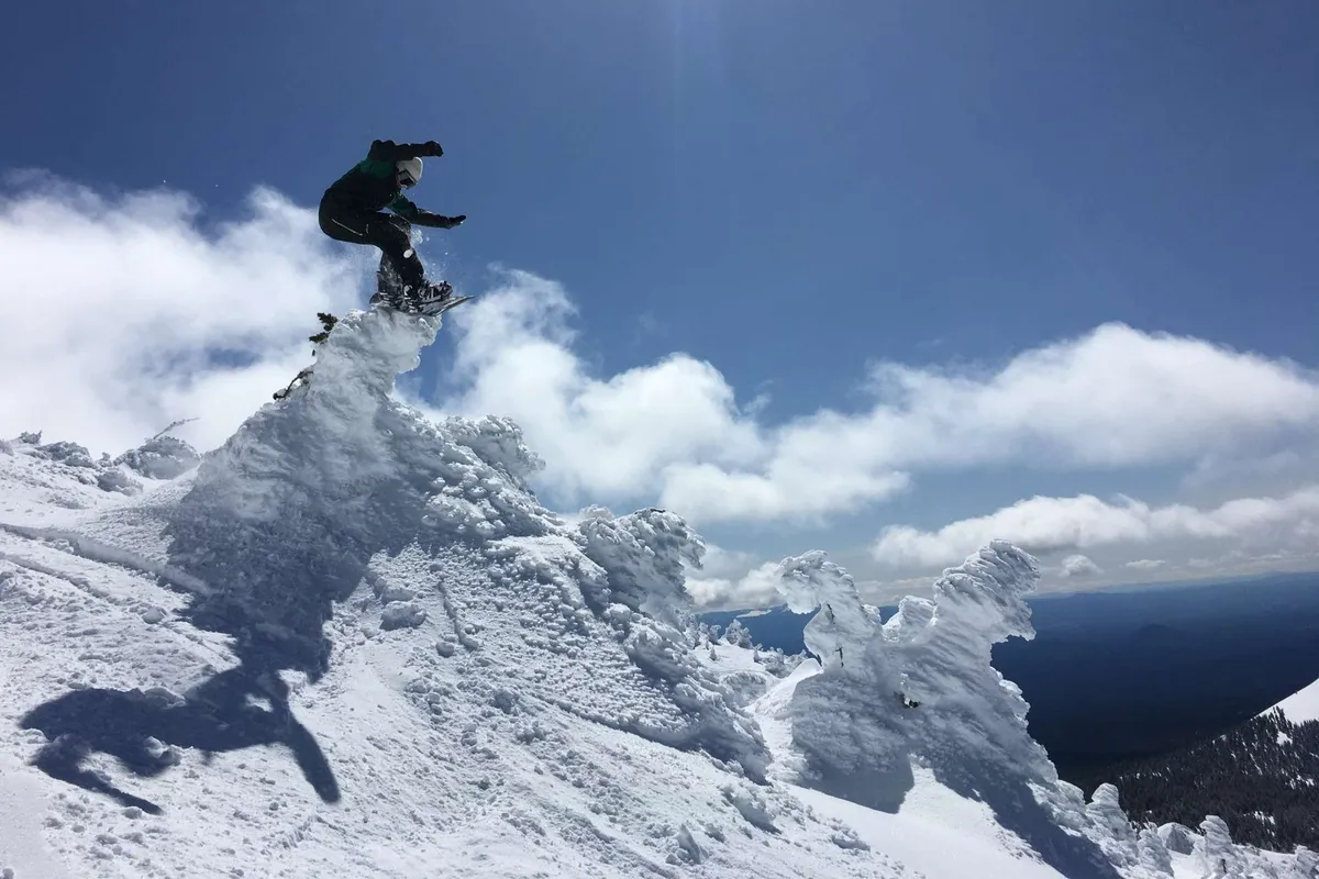 An exterior action shot capturing a snowboarder mid-air against the backdrop of a snow-covered mountain peak and dramatic sky. The surrounding landscape highlights the natural beauty and adventure opportunities in the Bend area.