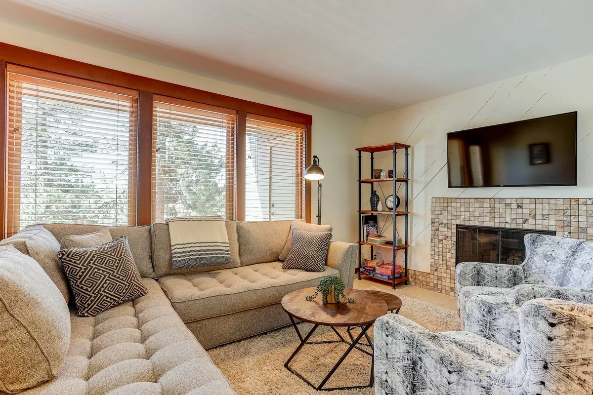 The image showcases a stylish, inviting living room featuring a plush sectional sofa, a cozy fireplace, a modern coffee table, and a bookshelf with thoughtful decorative accents. Morning sunlight streams in, illuminating the space and creating a warm, welcoming atmosphere perfect for relaxation or gathering with friends and family.