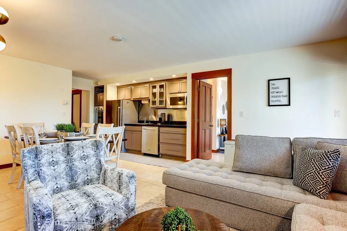 This image captures the heart of the open-concept space, featuring a comfortable gray sofa, a textured armchair, a dining table with chairs, and a well-equipped kitchen in the background. The space has a relaxed, welcoming mood, ideal for family gatherings and entertaining. The kitchen area is visible, showcasing modern finishes and open sightlines.