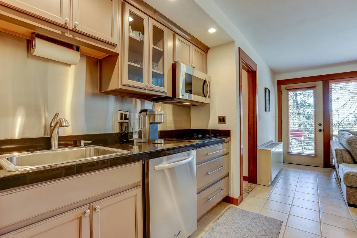 A bright, modern kitchen scene focused on stainless steel appliances, a kitchen sink, cabinetry, and a door leading outside. The kitchen is both functional and stylish, with granite countertops and high-quality finishes apparent. The open door suggests easy access for pets and outdoor flow.