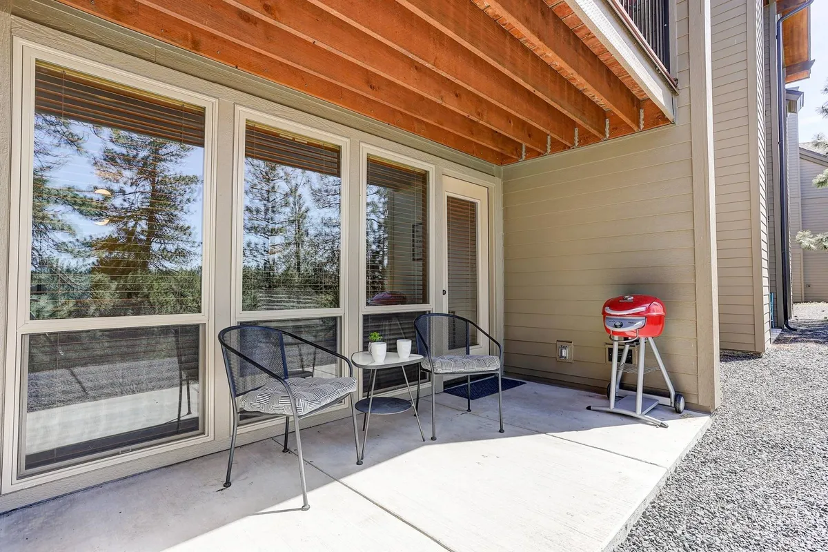 A cozy private patio space featuring two chairs and a small table, set against large windows with the backdrop of tall pine trees. A grill is also present, suggesting opportunities for outdoor cooking and relaxed gatherings. The setting is calm and intimate, inviting residents to enjoy morning coffee or social time amidst nature.