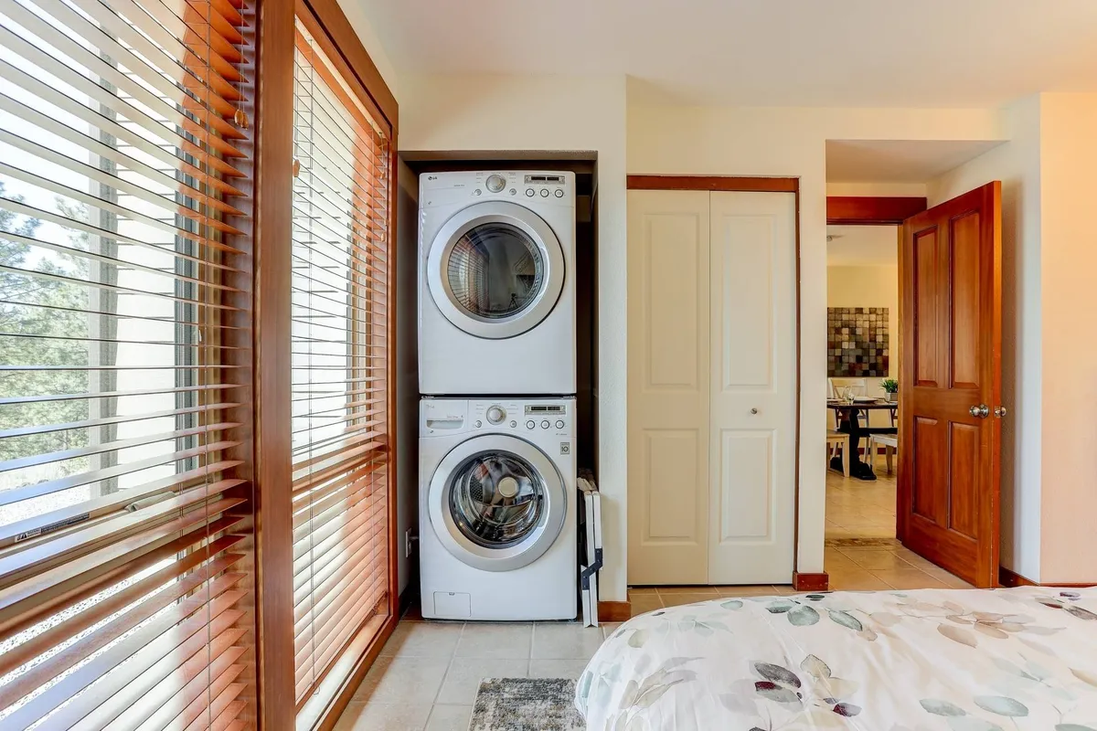 A functional, homey bedroom with a bed featuring floral bedding, adjacent to a window with blinds and a stacked washer and dryer in view. The abundance of natural light and practical amenities suggest comfort and convenience for guests and their pets.