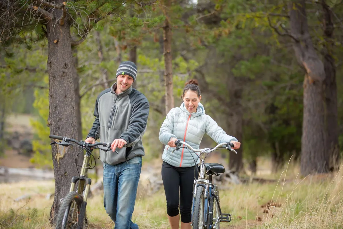 This lively exterior photo shows two people walking with bicycles across grassy terrain, set against a backdrop of tall trees. The atmosphere is joyful and active, suggesting family biking, picnics, and adventures within the resort or the nearby area.