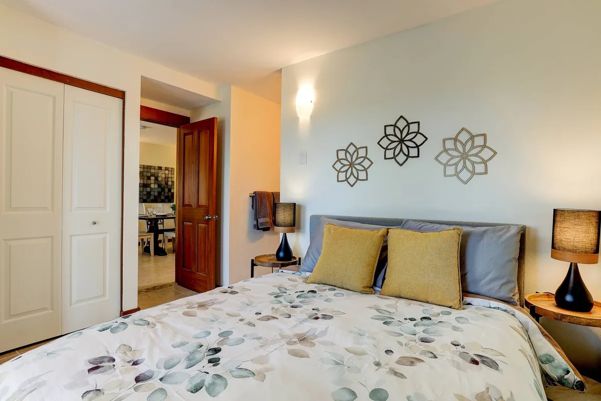 A serene bedroom featuring a bed with floral bedding, decorative wall art above the headboard, and matching nightstands with softly glowing lamps. Natural light streams in through a window, brightening the tranquil setting.