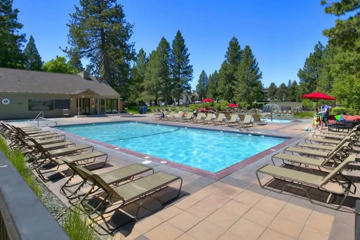 A lively exterior shot of the resort's heated swimming pool surrounded by lounge chairs, umbrellas, and a community building. Families are seen swimming, sunbathing, playing games, and barbecuing, with mature trees framing the scene.