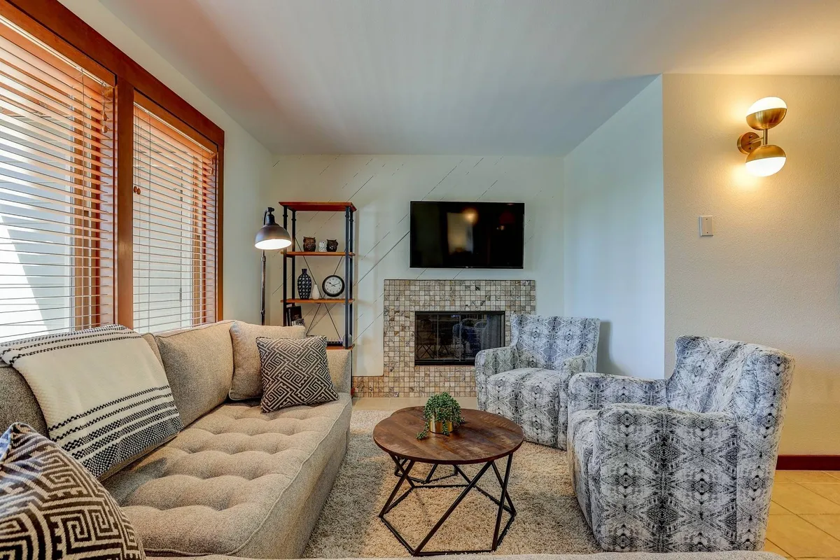 A cozy living room scene centered around a sofa, a fireplace, a coffee table, and shelves with decor. The mood is comfortable and inviting, with cues for relaxing by the fireplace or sharing a movie night. Expansive windows are present, hinting at natural views and outdoor connections.