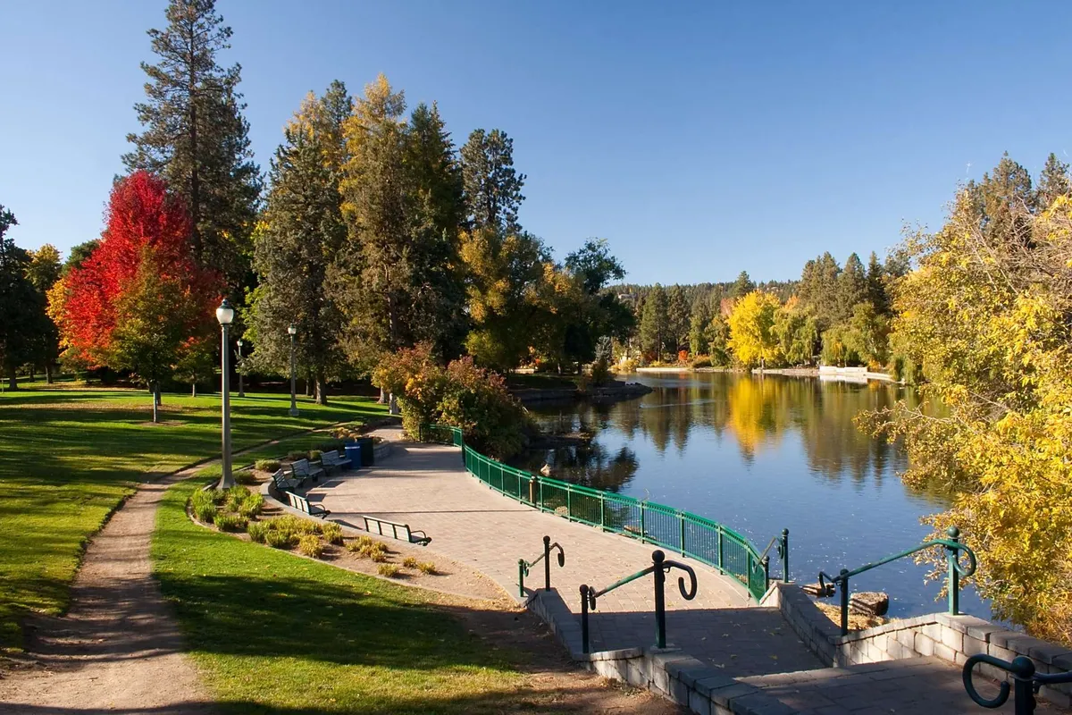 A peaceful riverside scene with a calm river, colorful trees, a walking path, park benches, and a green railing. The area looks inviting for a stroll, bird watching, or quiet reflection, perfectly embodying riverside living and easy nature access.